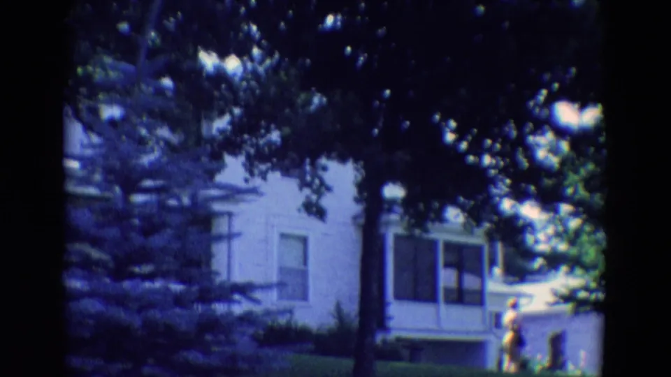 1969 Diller, Nebraska home-movie frame with a white house behind dense tree shade and a person near the yard edge.