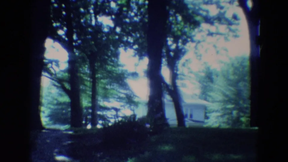 1969 Diller, Nebraska 8mm frame looking through trees across grass toward a low pale building in the background.