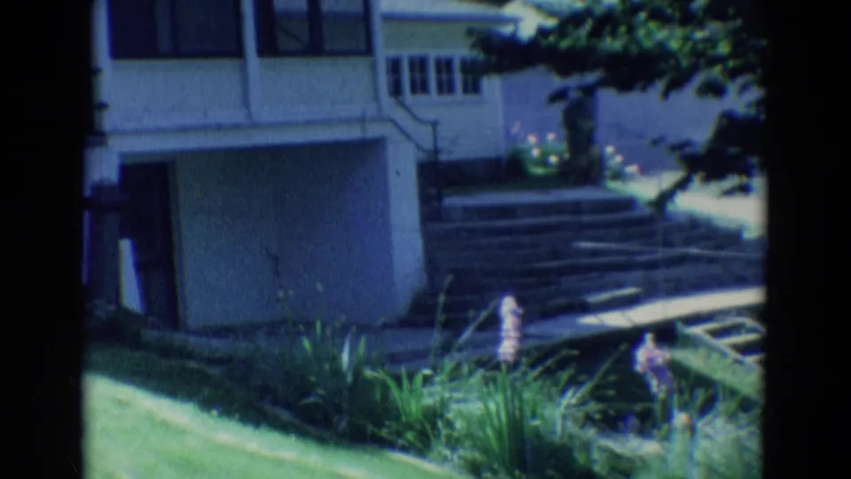 1969 Diller, Nebraska frame showing a yellow-sided house exterior with windows, garage door, steps, lawn, and garden flowers.