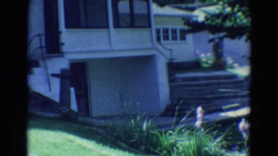 1969 Diller, Nebraska home-movie frame showing a white house exterior, front steps, lawn, and low garden plants.