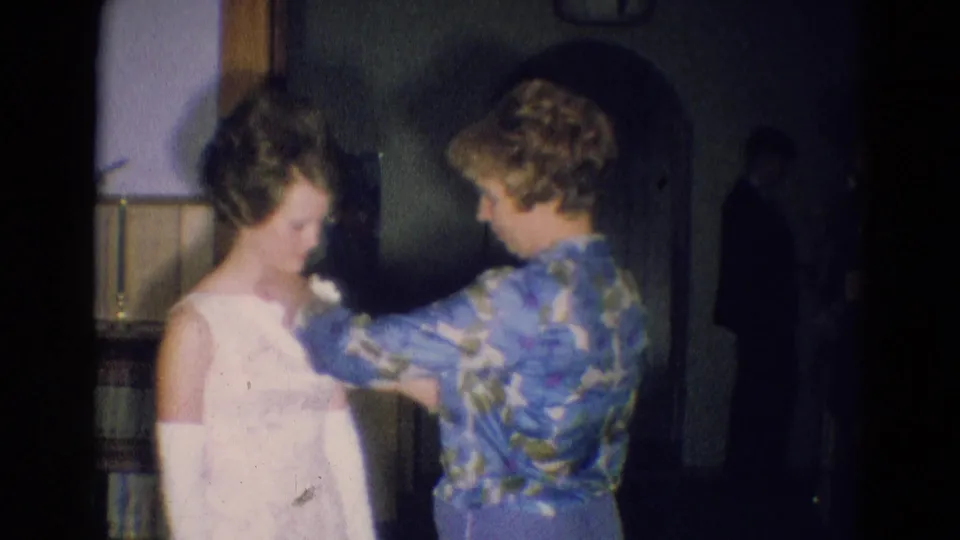 1969 Diller, Nebraska interior frame showing a woman pinning a corsage onto a young woman's white formal dress sleeve.