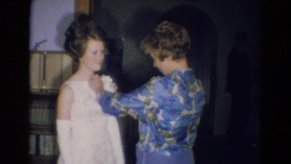 1969 Diller, Nebraska interior frame with a woman in a floral blouse adjusting a corsage on a white formal dress.