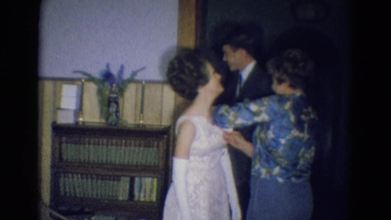 Young woman in a white dress stands as another woman adjusts her clothing while a suited man stands nearby.