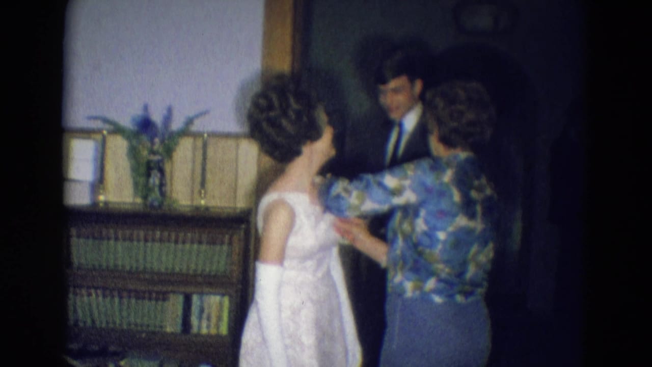 The white-dressed young woman faces a man in a suit as another woman continues adjusting the dress inside a Diller home setting.