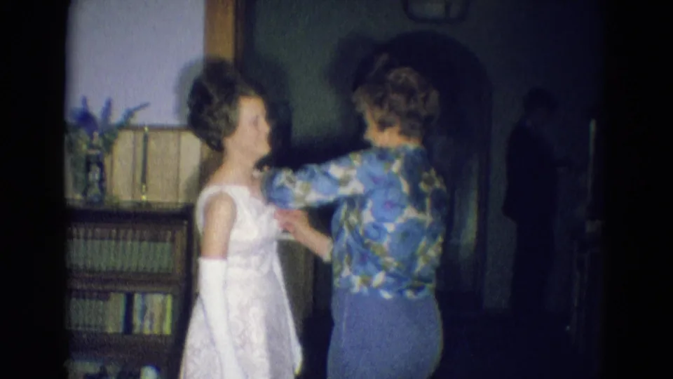 A woman in a blue floral blouse works on the front of a young woman's white formal dress in a dim 1969 Nebraska home interior.
