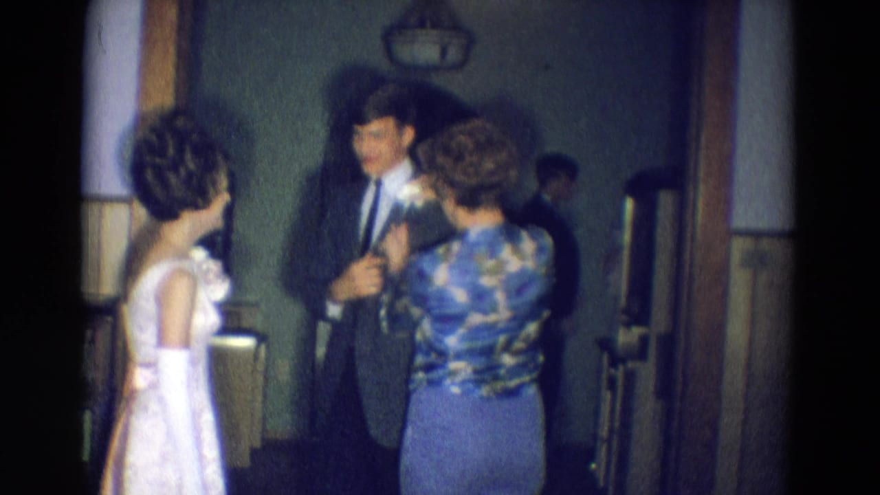 Young man in a dark suit stands as a woman reaches toward his lapel while a young woman in a light dress watches.