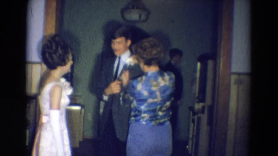 Diller, Nebraska 1969 home-movie frame showing a young man in a suit before a prom-like event as a woman prepares to pin a flower.