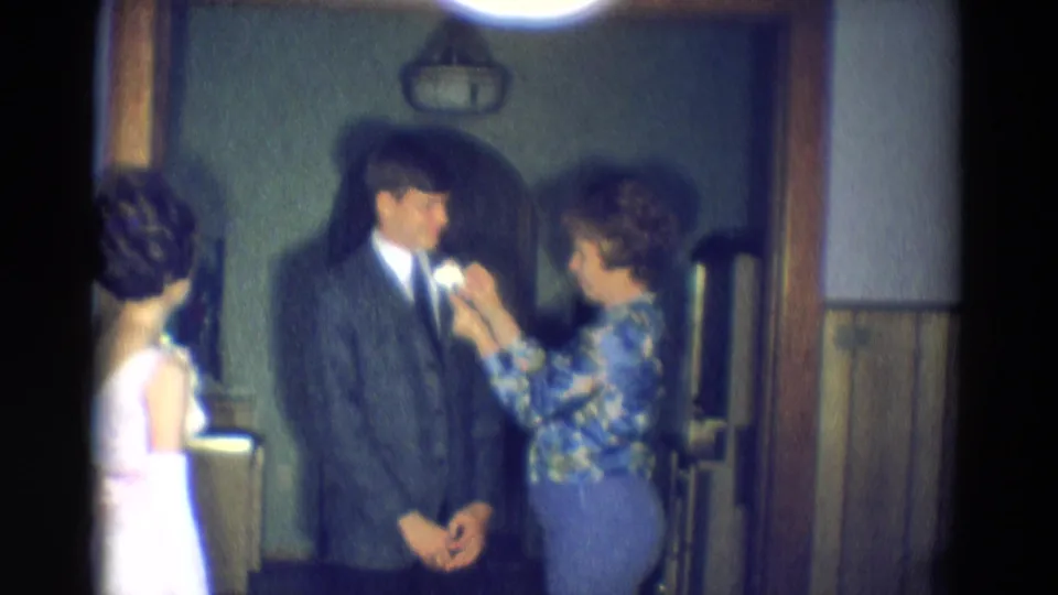Diller, Nebraska 1969 home-movie still with a woman adjusting a white lapel flower on a suited young man before a formal event.