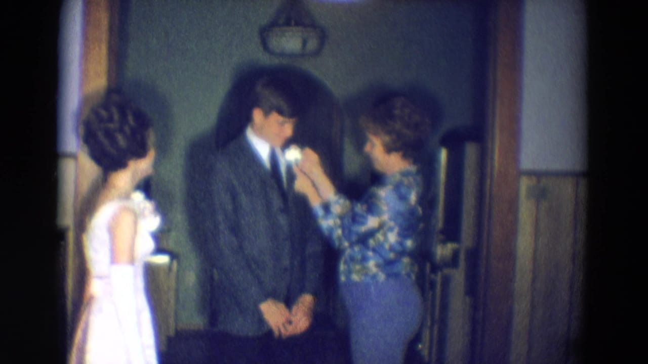 Woman pins a boutonniere on a young man in a suit while a young woman in a white dress watches.