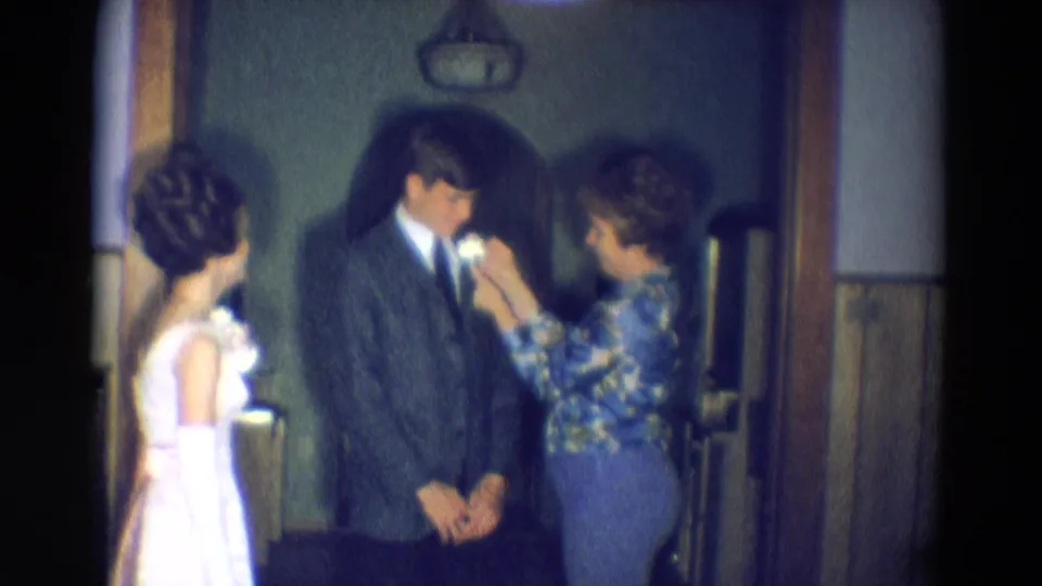 In Diller, Nebraska, 1969 home-movie footage, a woman adjusts a boutonniere on a suited young man as a young woman in a white formal dress stands nearby.
