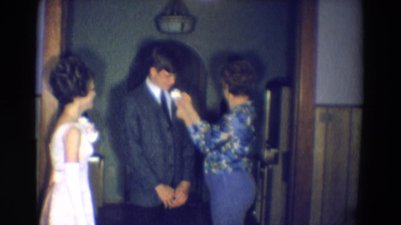Young man looks down as a woman pins a boutonniere, with a young woman in white at left.