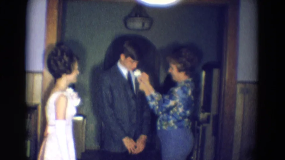 In this 8mm frame from Diller, Nebraska, a woman continues fastening a boutonniere to a young man's suit while a young woman in a white dress stands beside them.
