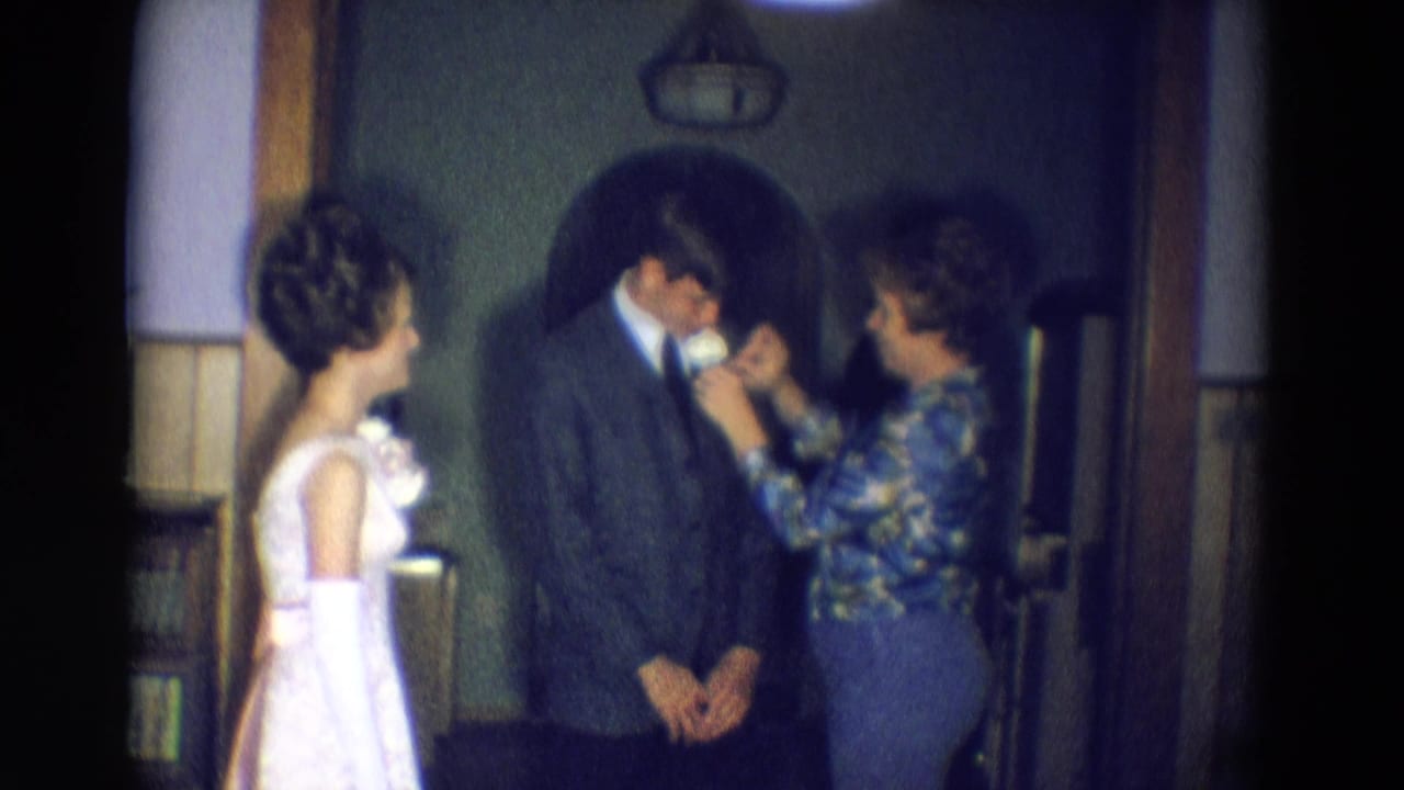 Adults in formal clothing stand in a wood-paneled hallway as a woman adjusts a man's tie.