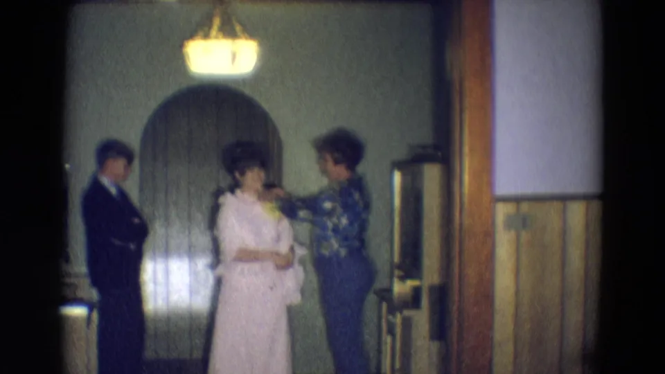 In 1969 Diller, Nebraska home-movie footage, a woman adjusts another woman's pale formal dress while a suited man waits nearby.