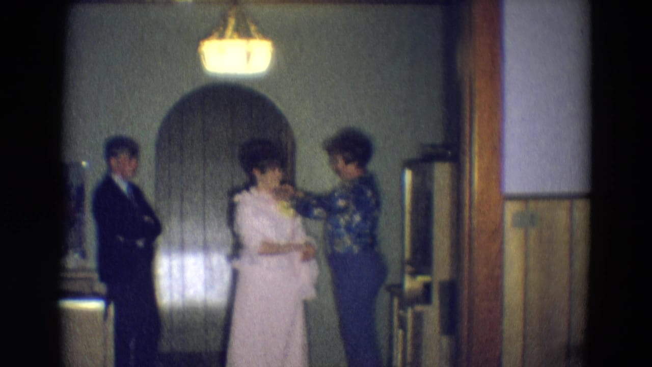 A woman adjusts another woman's dress while a suited man stands near the doorway.