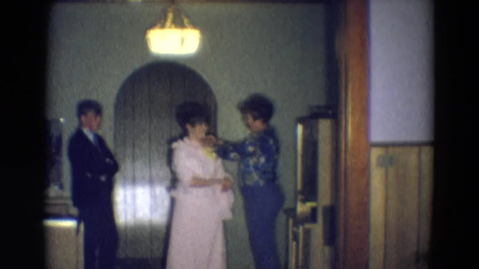 A floral-bloused woman tends to a pale gown as a man in a dark suit waits in the softly lit Diller, Nebraska interior.