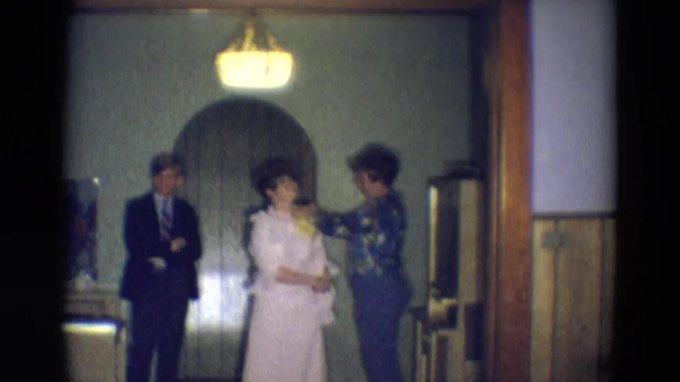 The man shifts slightly with arms crossed as the two women remain focused on the pale formal dress in the 1969 home-movie scene.