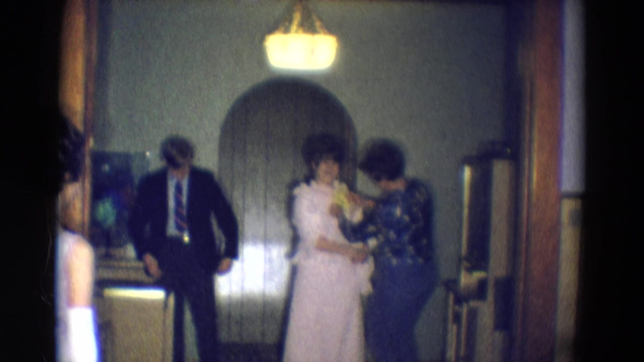 In a Diller, Nebraska 1969 home-movie frame, a woman in a white dress is adjusted by another woman as a man in a dark suit waits nearby.