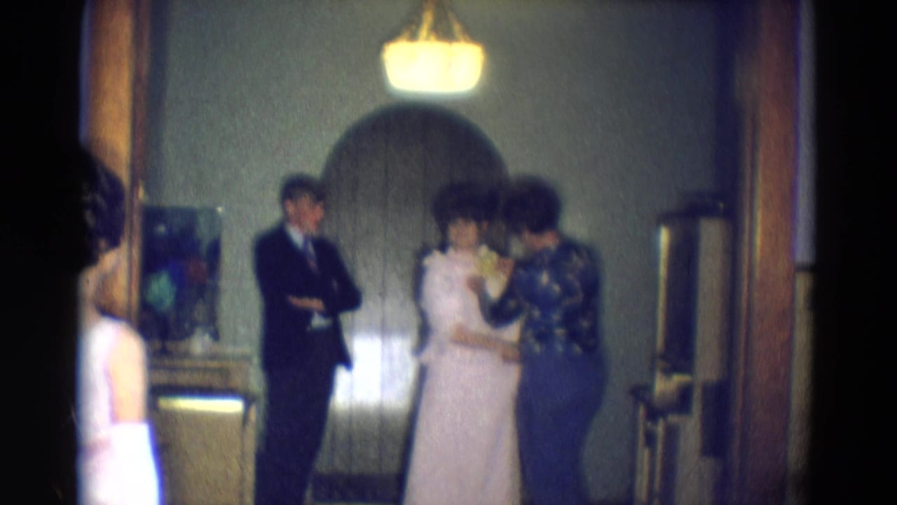 A suited man stands with arms crossed as a woman in a white dress is adjusted by another woman.