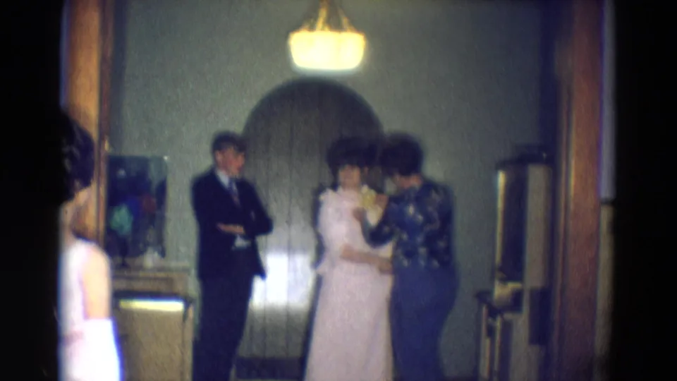 The dress fitting continues in the dim interior, with the suited man watching while the helper works near the front of the white dress.