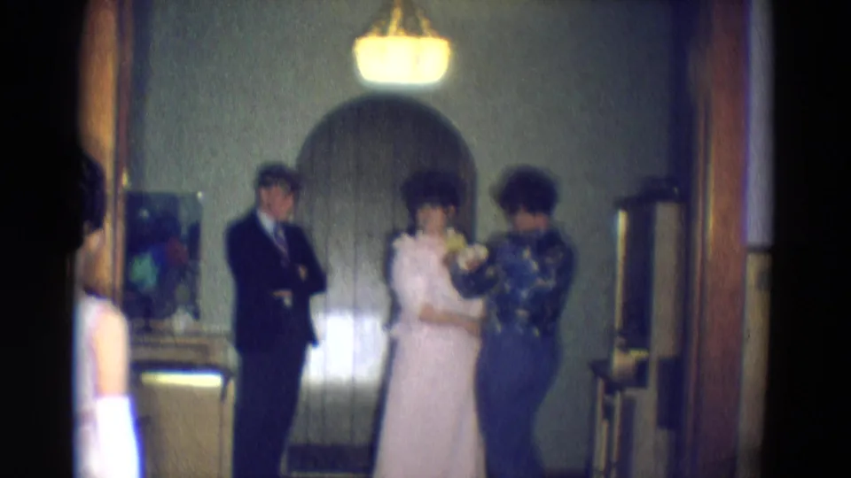 A softly focused 1969 8mm frame shows the woman in white standing for fitting while the man in a suit waits beside the arched doorway.