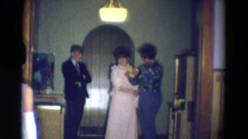 The final frame keeps the dress adjustment centered, with the helper facing the woman in white and the suited man watching in the interior.