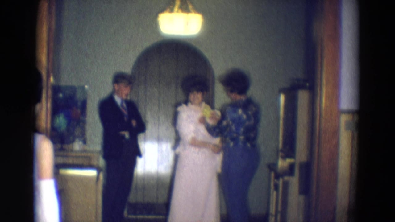 Bride and mother stand with flowers while a man in a dark suit watches indoors.