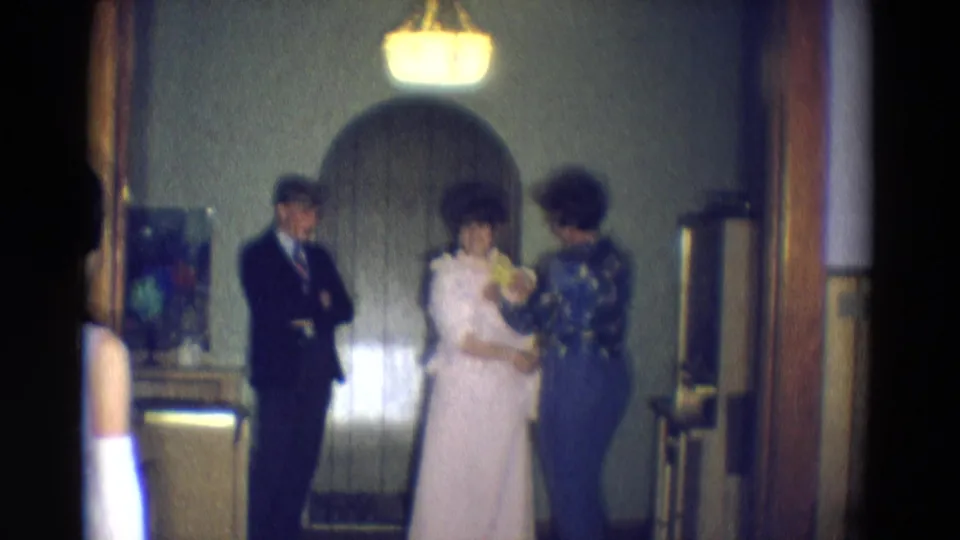 In Diller, Nebraska in 1969, a bride in white stands beside her mother receiving yellow flowers as a suited man watches inside a dim room.