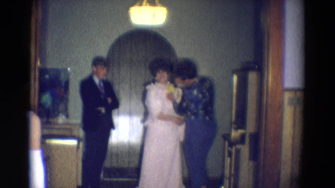 Bride and mother hold yellow flowers near an arched doorway with a suited man nearby.