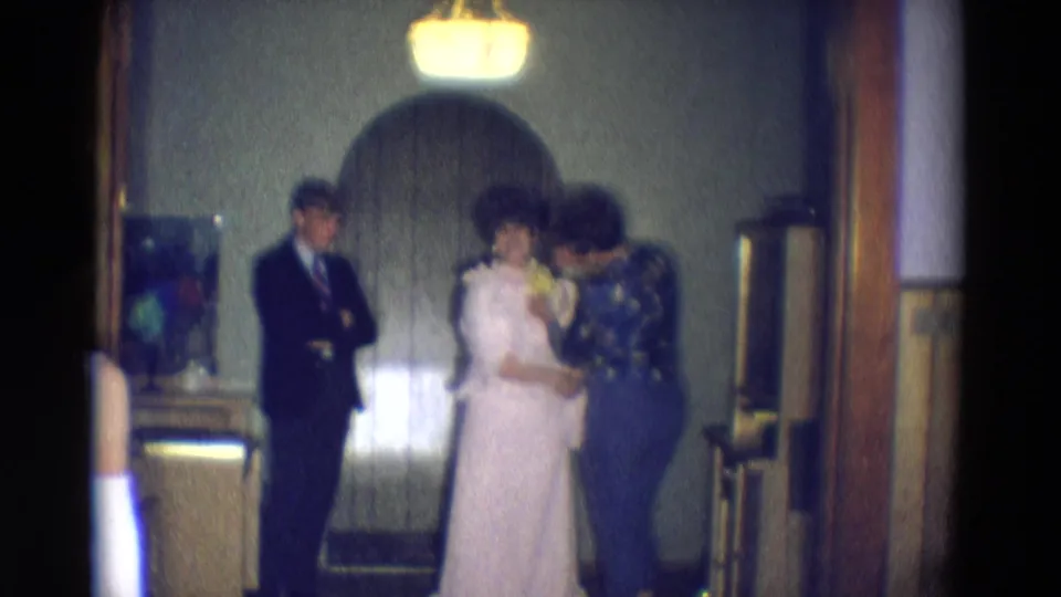 A 1969 home-movie frame shows the bride and her mother with yellow flowers near an arched doorway in a private Diller, Nebraska interior.