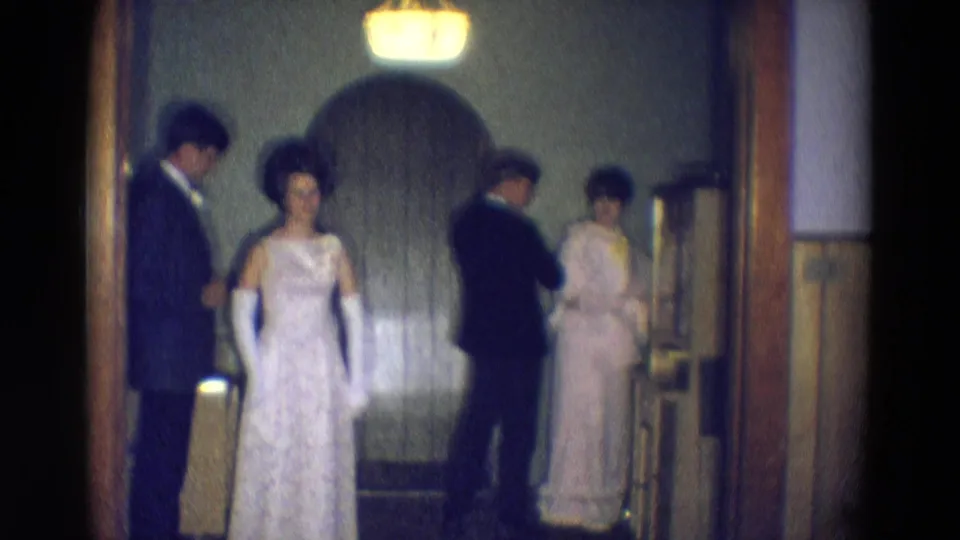 The bride faces the camera in her white lace gown while relatives in suits and formal dresses stand nearby in the 1969 Nebraska wedding room.