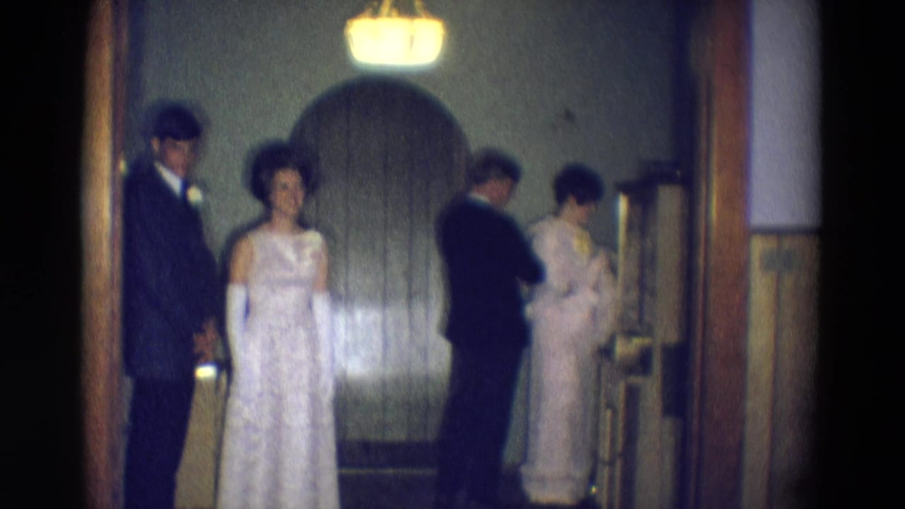 Two formally dressed couples stand in a dim hallway near a wooden doorway.