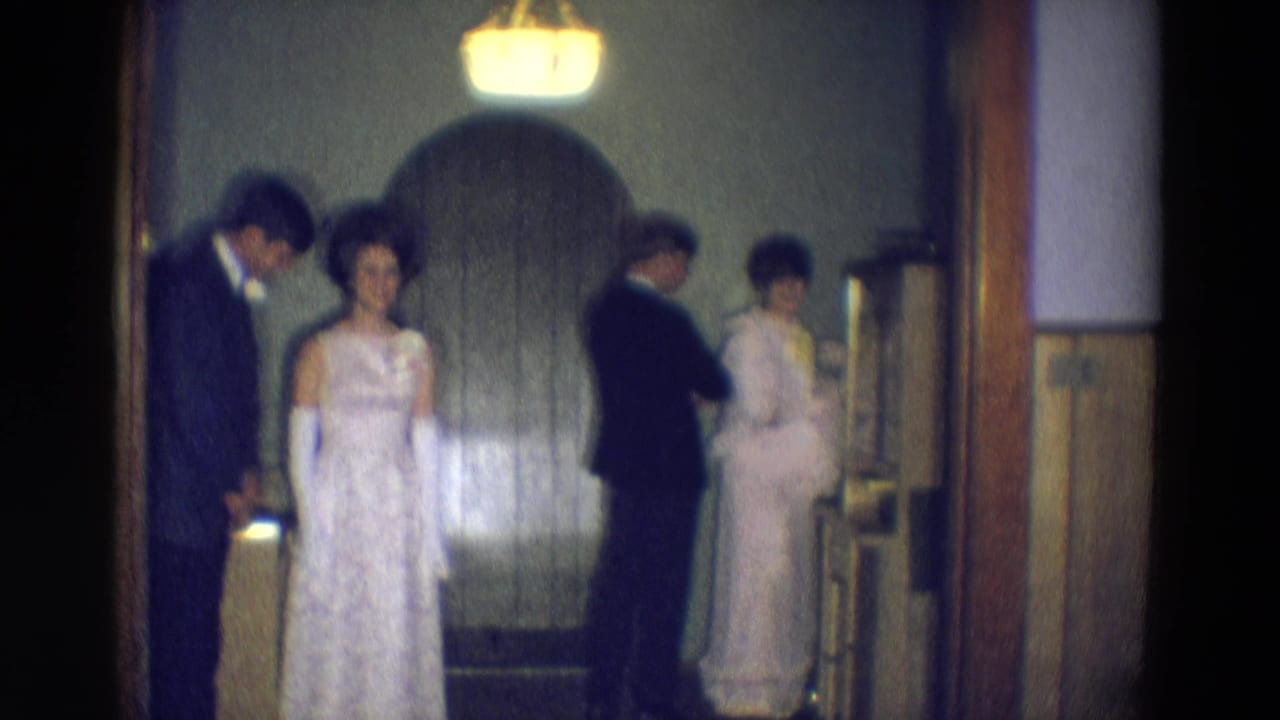 A woman in a white gown faces the camera while others stand near a doorway.