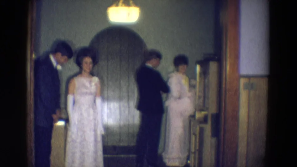 A woman in a white formal gown and gloves faces the camera as other adults in suits and light dresses stand nearby in the 1969 Diller scene.