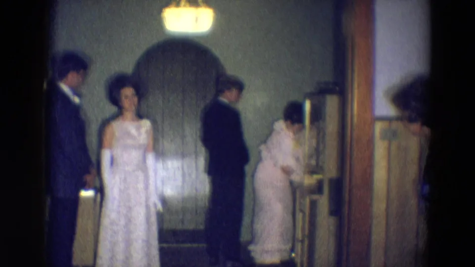During the Diller, Nebraska 1969 home movie, formal guests wait as a woman in a light dress bends near the doorway or wall fixture.