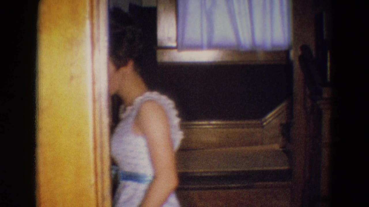 In a 1969 Diller, Nebraska home interior, a young woman in a pale formal dress passes near a doorway beside a staircase.