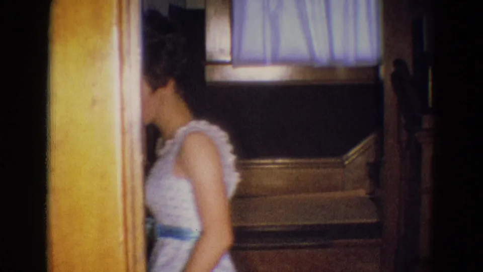 In a 1969 Diller, Nebraska home interior, a young woman in a pale formal dress passes near a doorway beside a staircase.