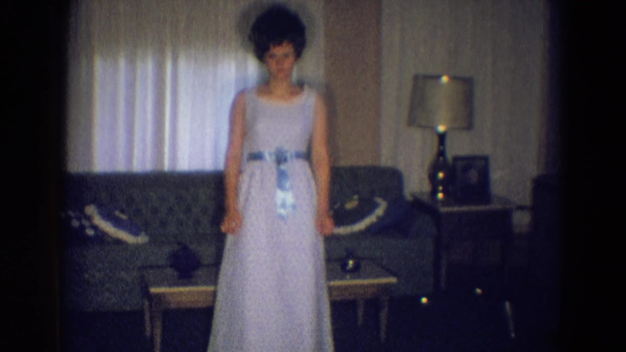 Young woman stands in a pale dress before a sofa and curtained window.