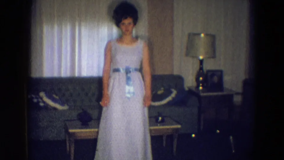The woman faces the camera in a pale formal dress, standing before a tufted sofa, coffee table, curtained window, and lamp in Diller, Nebraska.