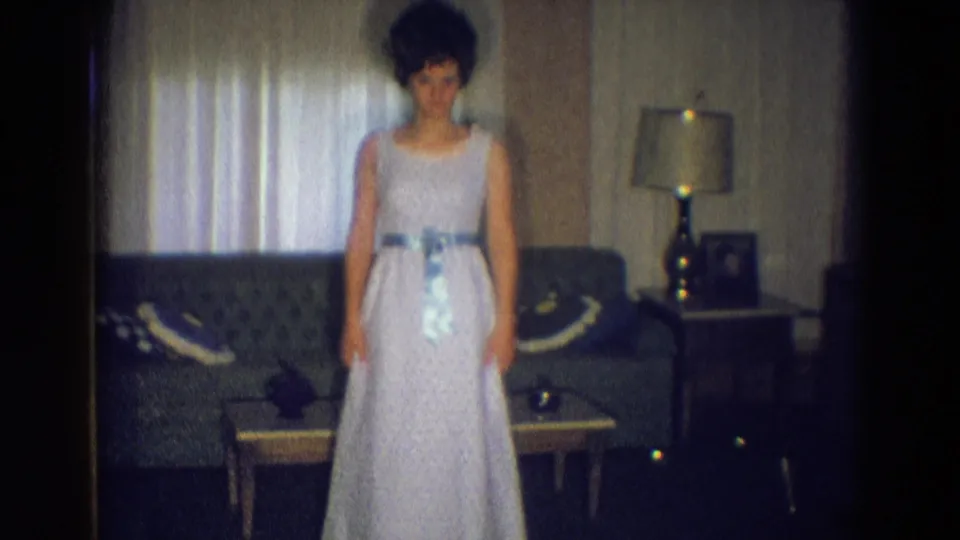 A young woman remains posed in a pale dress with a blue sash in a 1969 Diller, Nebraska living room with sofa, table, lamp, and curtains.