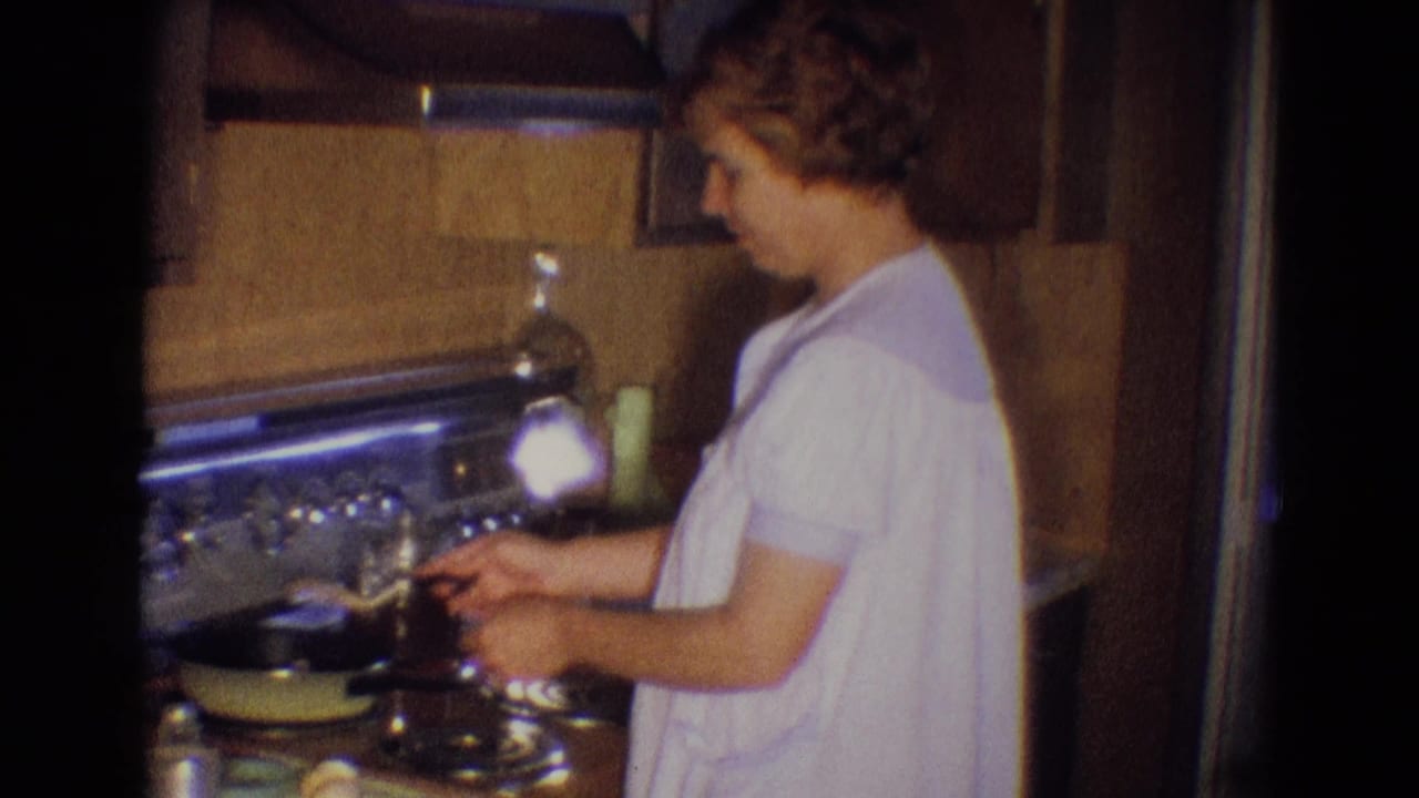 Woman in a white gown cooks at a stovetop in a wood-paneled kitchen.