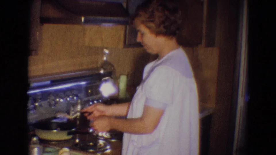 In 1969 Diller, Nebraska home-movie footage, a woman in a white gown stands at a stove while cooking eggs in a kitchen.