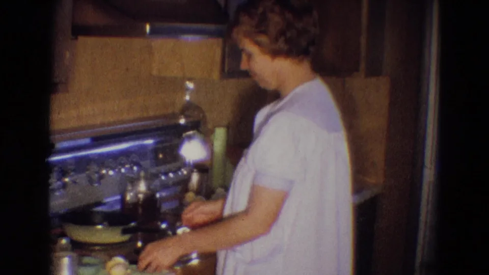 A woman in a white gown works at a kitchen stove in Diller, Nebraska, with cookware and wood cabinetry visible around her.