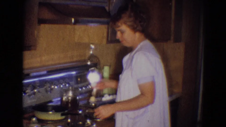 The woman holds an egg beside the stove in this 1969 Diller, Nebraska kitchen scene, with pans and cabinetry in view.