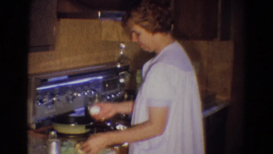 A woman in a white gown holds an egg over the stove in a private Diller, Nebraska kitchen, recorded on 8mm home-movie film.