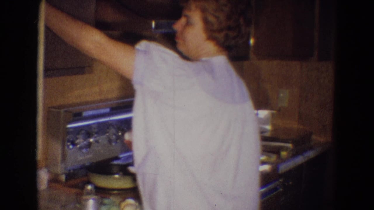 In a 1969 Diller, Nebraska home kitchen, a woman in a light nightgown reaches toward an upper cabinet near a metal stove.