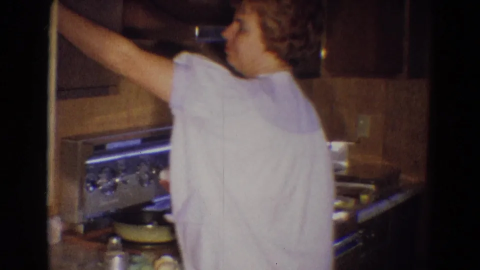 In a 1969 Diller, Nebraska home kitchen, a woman in a light nightgown reaches toward an upper cabinet near a metal stove.