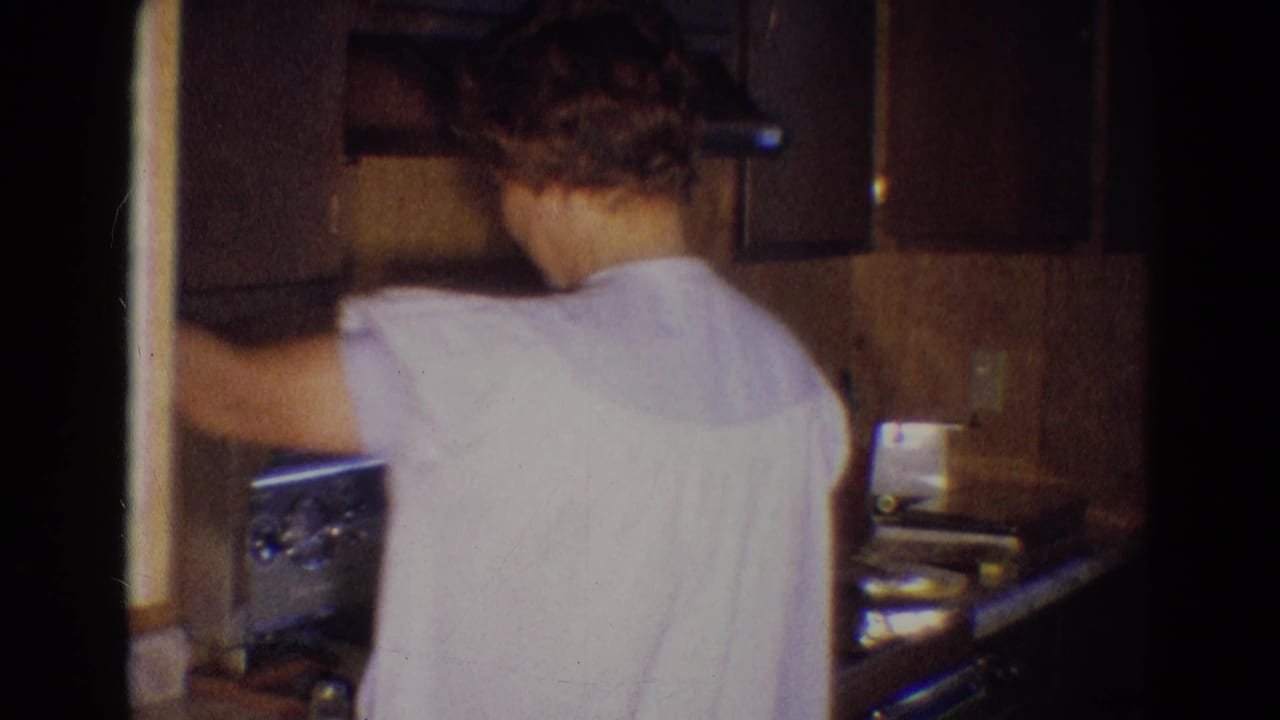 Woman stands with her back to the camera at a stove in a wood-paneled kitchen.