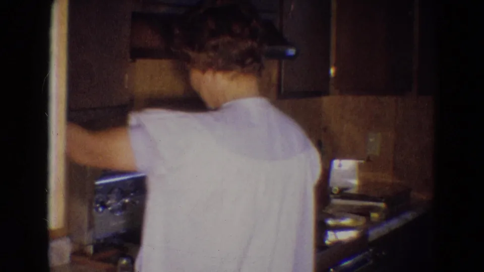 The woman faces the stove in the Diller, Nebraska kitchen, with dark wood cabinets and counter surfaces around her.