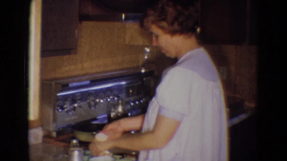 The woman works over a pan on the stove, appearing to crack or handle an egg during breakfast preparation.